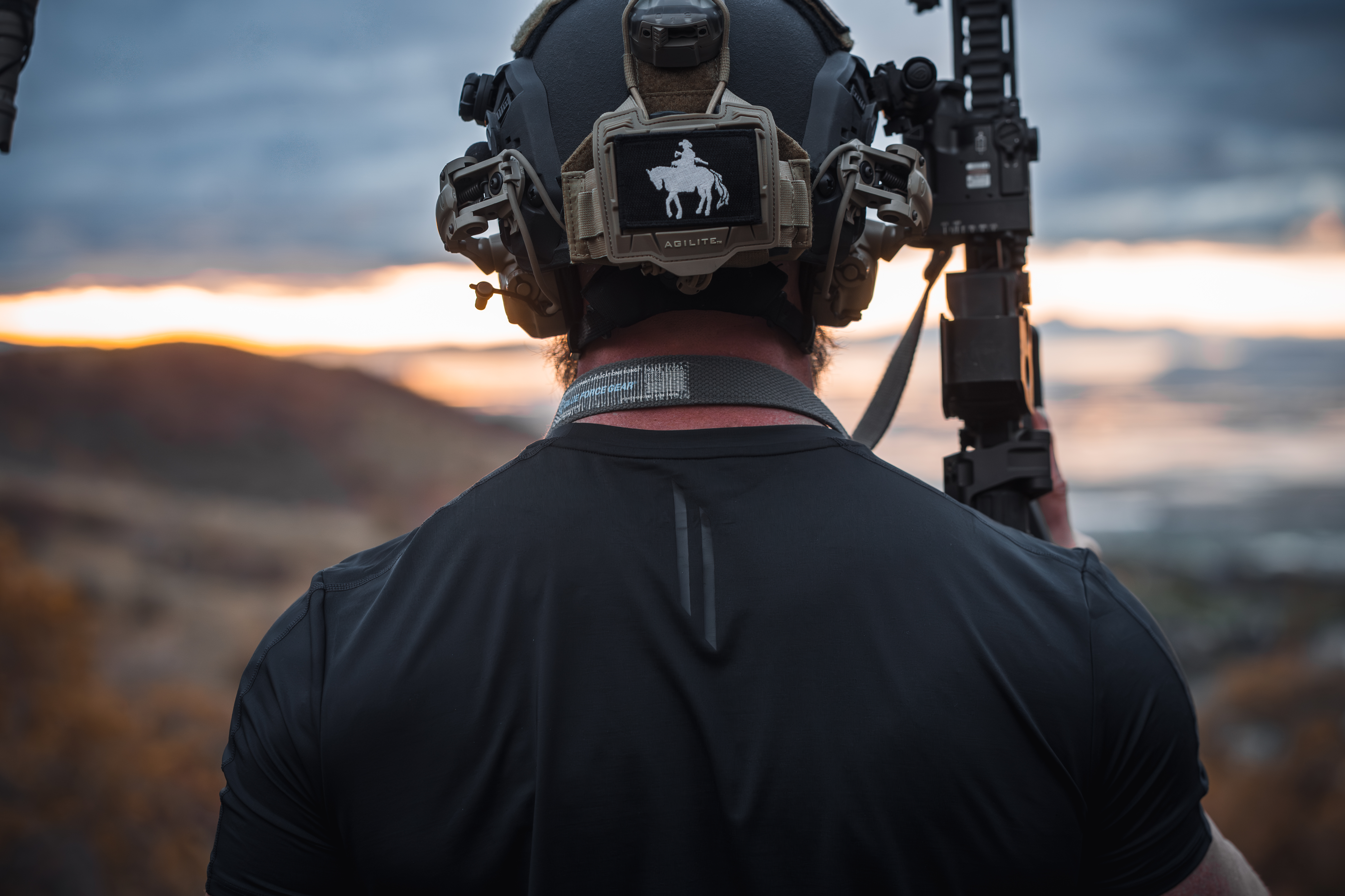 LINEAR Clothing model with his back to the camera wearing Baseline Series shirt wearing a tactical helmet and holding a rifle with the sunset behind him